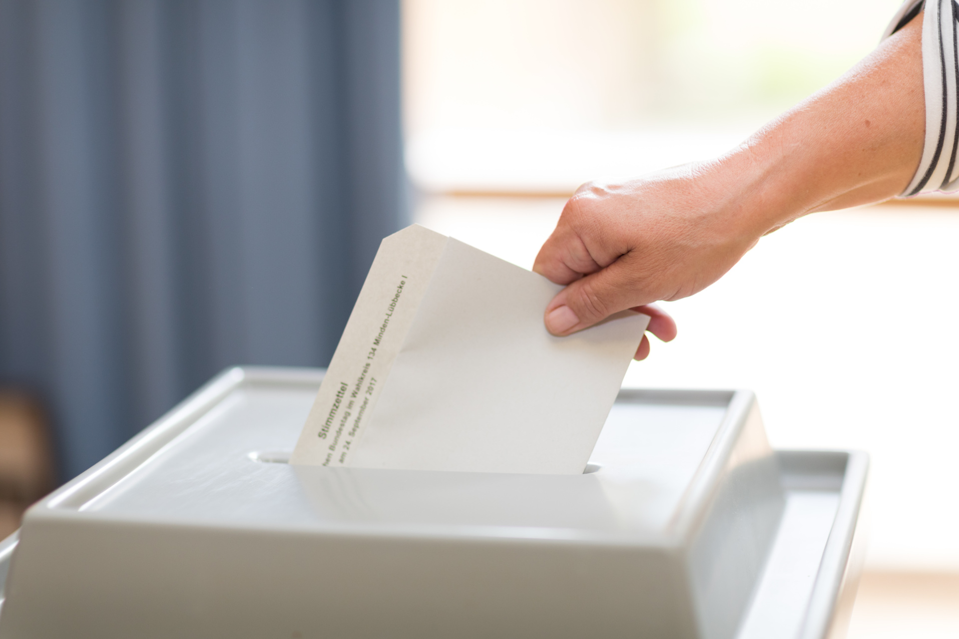 Eine Hand wirft einen weißen Briefumschlag mit der Aufschrift „Stimmzettel“ in den Schlitz einer hellgrauen Wahlurne. Die Person trägt ein gestreiftes Oberteil.