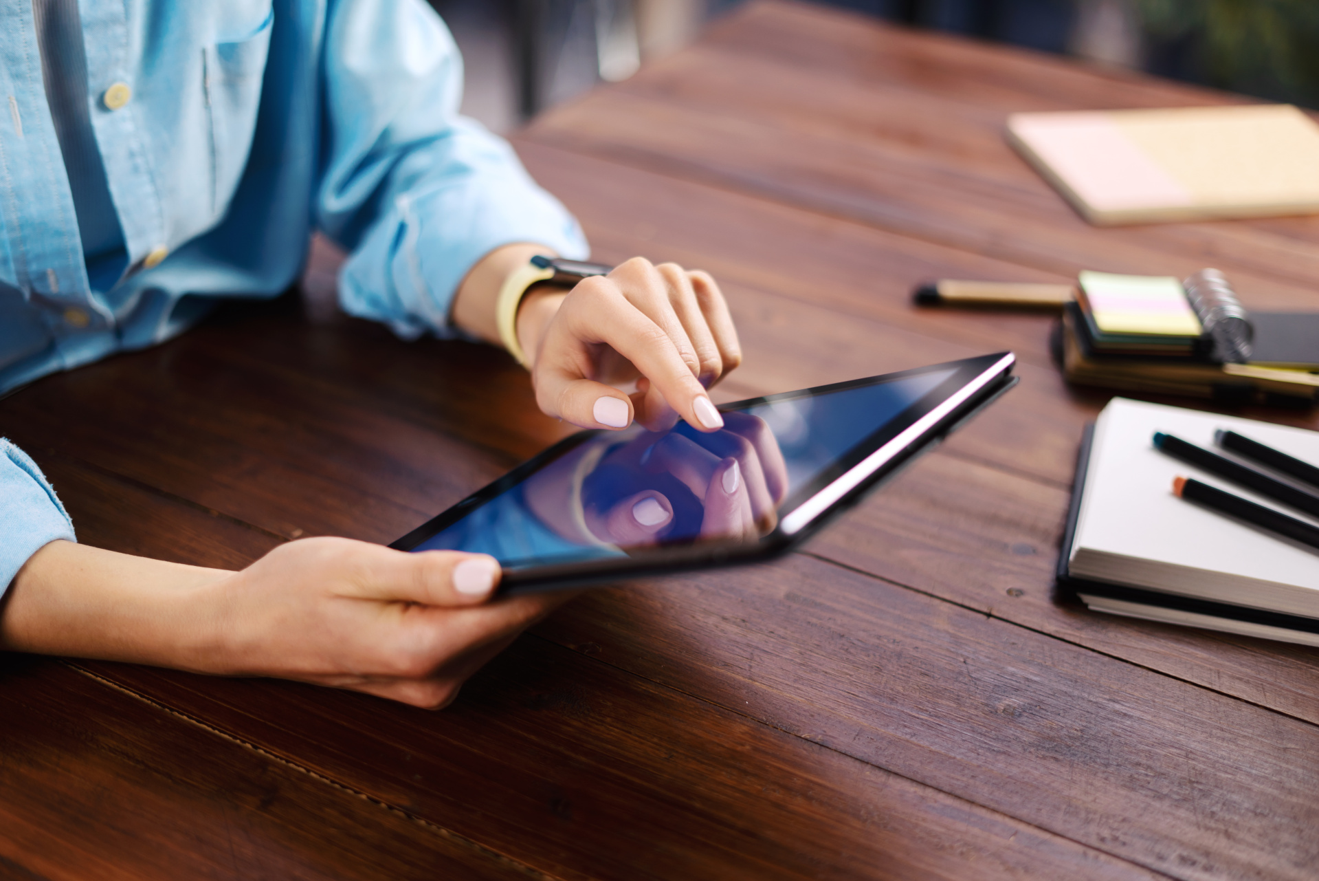 Eine Person in einem hellblauen Hemd bedient ein schwarzes Tablet auf einem dunklen Holztisch. Mit dem rechten Zeigefinger berührt sie das Display, während die linke Hand das Tablet stützt. Im rechten Bildbereich liegen Notizbücher und Stifte auf dem Tisch. Der Hintergrund ist hell und unscharf.