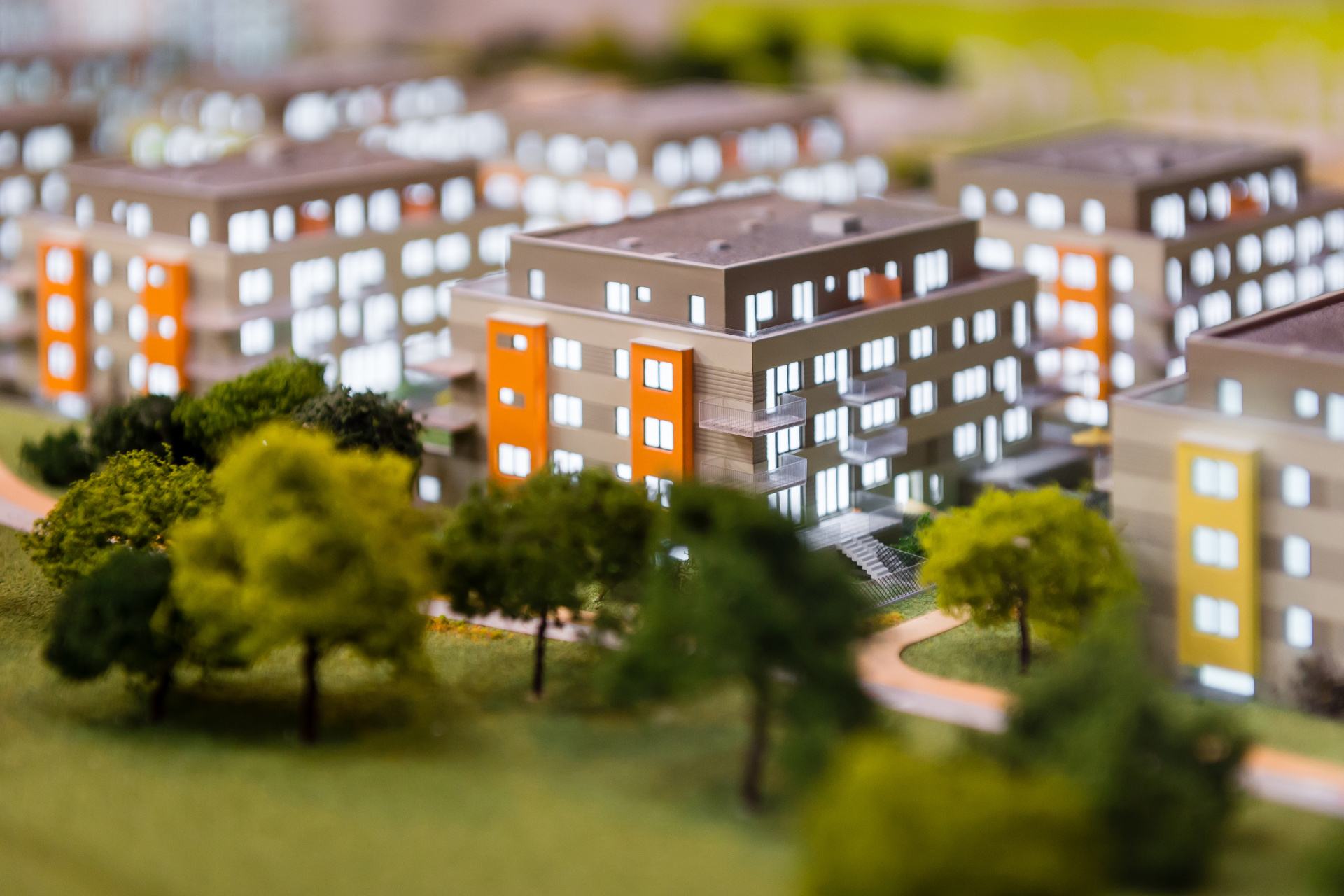 Architekturmodell von Gebäuden Ein Architekturmodell mit mehreren modernen, mehrstöckigen Gebäuden, die graue Fassaden und orangefarbene Akzente haben. Die Gebäude sind von kleinen Modellbäumen und Grünflächen umgeben.