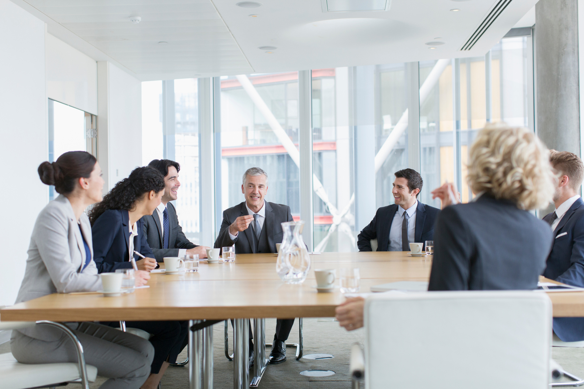 Eine Gruppe von Personen in Business-Kleidung sitzt an einem großen Holztisch in einem hellen, modernen Konferenzraum mit großen Glasfenstern. Ein Mann in der Mitte spricht und gestikuliert mit der Hand, während die anderen Personen ihm aufmerksam zuhören.