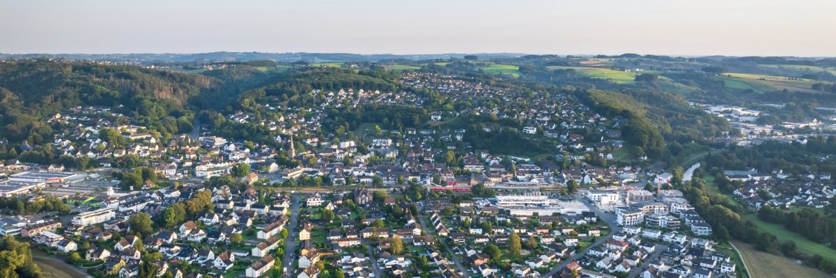 Eine weite Luftaufnahme der Stadt Overath, eingebettet in eine grüne, hügelige Landschaft. Man sieht eine dichte Besiedlung mit vielen Einfamilienhäusern mit dunklen Dächern und einigen größeren Geschäfts- und Wohngebäuden im Zentrum. Die Stadt ist von weiten Wäldern und Feldern umgeben. Im Vordergrund ist ein bewaldeter Hang zu sehen.
